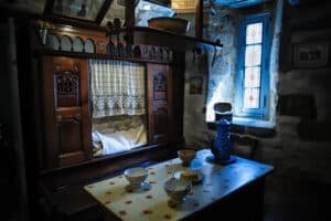 Lit clos breton en bois sculpté à portes à fuseaux et rosaces, rideau de dentelle, devant une table couverte d'une toile cirée avec bols et cafetière émaillée bleue, dans une maison en granit de Pont-Croix (Finistère)