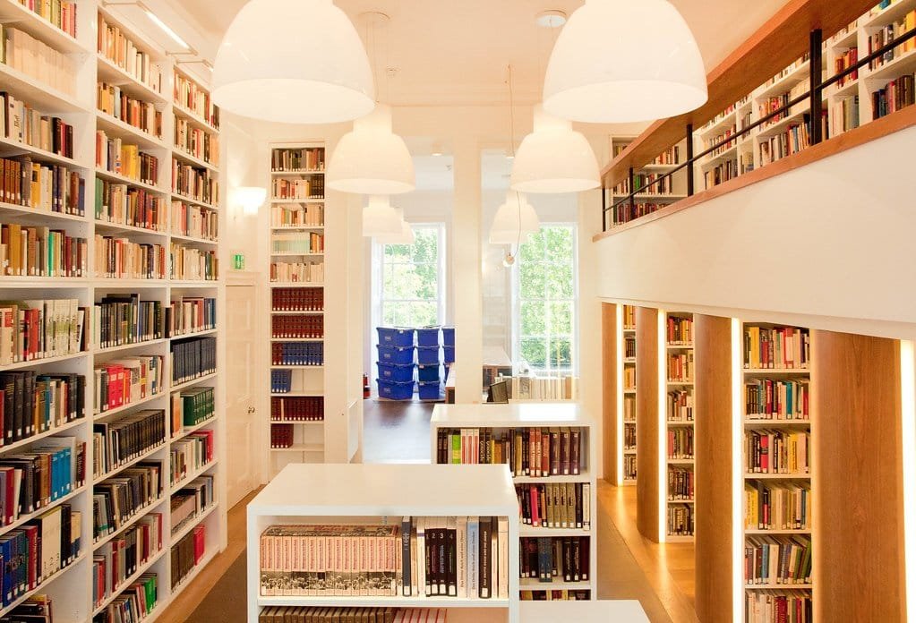 Salle de bibliothèque avec rayonnages blancs sur mesure, mezzanine, meubles bas et piliers habillés de bois