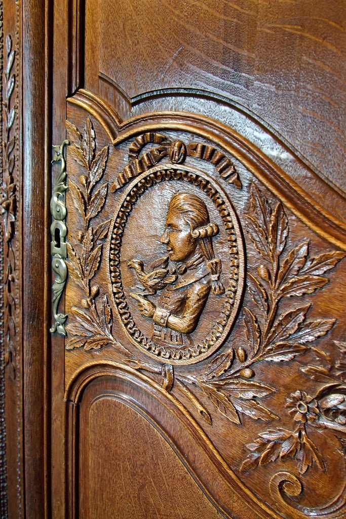 Détail d'une porte d'armoire de mariage normande en chêne : médaillon sculpté d'un jeune homme en perruque tenant une colombe, entouré de rameaux d'olivier et de fleurs, entrée de serrure en laiton
