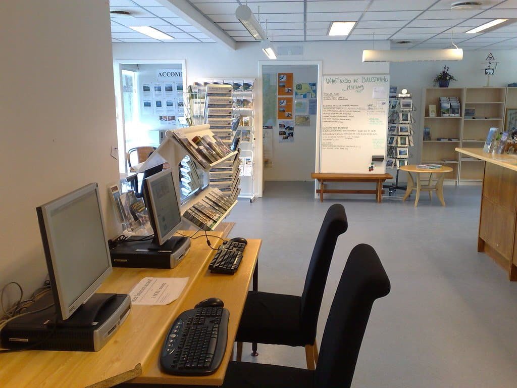 Intérieur d'un office de tourisme avec postes informatiques, présentoirs à brochures, étagères et comptoir en bois