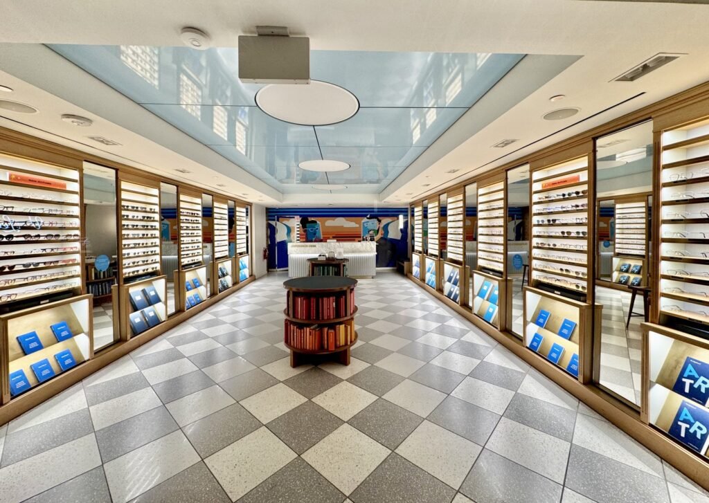 Magasin d'optique avec présentoirs muraux à lunettes éclairés en bois clair, sol en damier et comptoir au fond