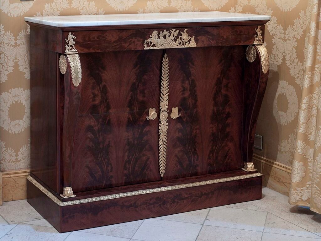 Commode à deux vantaux en acajou flammé, dessus de marbre blanc, montants en console galbée, ornements de bronze doré (char, feuillages, papillons), vers 1820